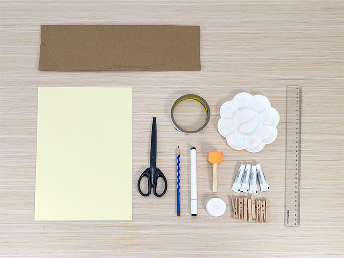 An assortment of craft supplies lies neatly arranged on a light wood-grain table. The supplies include a piece of brown cardboard, a sheet of light yellow paper, a roll of clear tape, scissors, a pencil, a white marker, a small orange sponge brush, a flower-shaped paint palette, several small tubes of paint or glue, a small white container with a lid, a ruler, and a handful of wooden clothespins. This flat lay image suggests preparation for a craft or art project.