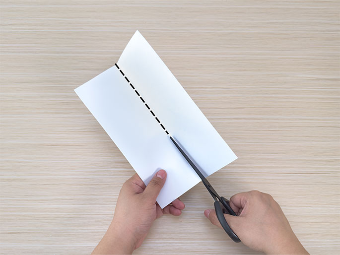 Two hands are shown cutting a piece of white paper with scissors on a light wood-grain background. The paper is folded in half and a dashed line is visible down the center fold, guiding the cut. The hands hold the paper and scissors, actively in the process of cutting along the line.