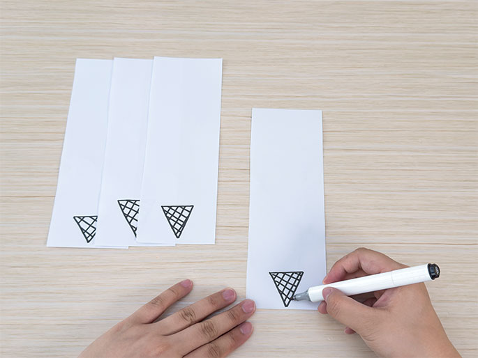 On a light wood-grain surface, several strips of white paper are laid out. Three of the strips already have a simple, black line drawing of an ice cream cone at the bottom. A hand holding a white marker is in the process of drawing the same ice cream cone design on the bottom of a fourth strip of paper. This image suggests a crafting activity, possibly making bookmarks or decorative items.