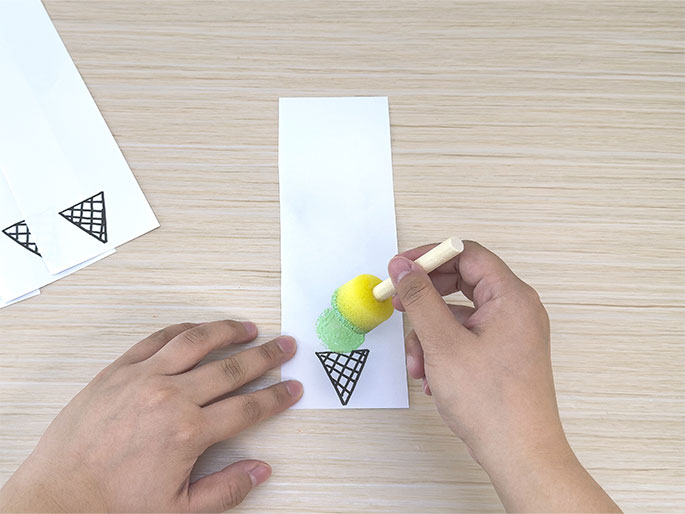 On a light wooden surface, a hand uses a small, round sponge brush to apply light green paint or ink to a drawing of an ice cream cone on a strip of white paper. The cone outline is drawn in black ink, and the green color is being added to represent the ice cream. In the background, other similar strips of paper with ice cream cone outlines are visible, suggesting a crafting project in progress.