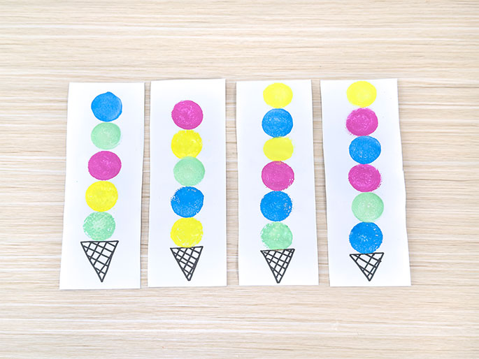 Four identical pieces of white paper are arranged on a light wood-grain surface. Each paper features a hand-drawn ice cream cone at the bottom, topped with a vertical stack of colorful circles representing ice cream scoops. The colors and order of the scoops vary on each paper. From left to right, the scoop colors are: blue, green, pink, yellow, light green; pink, yellow, light green, blue, yellow; yellow, yellow, pink, blue, light green; and yellow, pink, blue, light green, blue. These papers likely represent a crafting activity or a game involving color and pattern recognition.