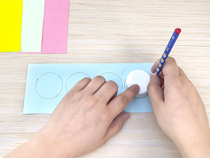 Two hands are shown tracing circles onto light blue paper using a small, white plastic lid as a template and a pencil. Several lightly drawn circles are already visible on the paper to the left of the hands. In the top left corner, sheets of yellow, light green, and pink paper suggest these colors will be used for the circles once they are cut out. This image depicts a crafting activity, likely involving creating circular cutouts for a project.
