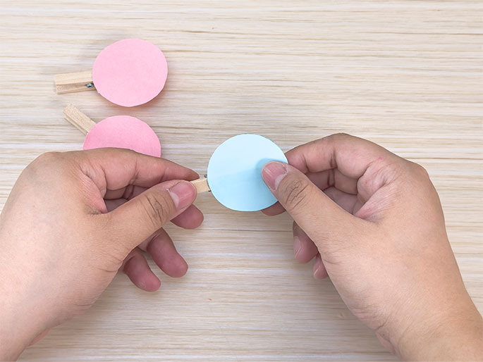 Two hands are shown attaching a light blue paper circle to a small wooden clothespin. Two other clothespins with pink paper circles attached are visible in the background on the light wood-grain surface. This image suggests a crafting activity, possibly making paper ornaments or decorations using clothespins.