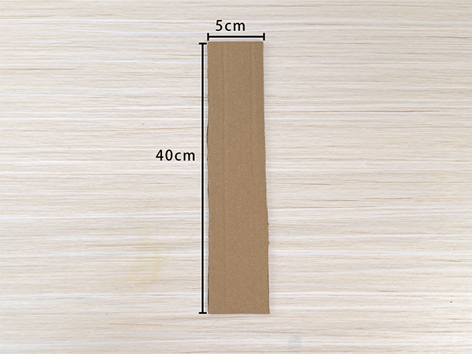 A long, narrow strip of brown cardboard lies on a light wood-grain surface. Lines indicating measurements are drawn alongside the cardboard, showing it to be 40cm long and 5cm wide. This image likely depicts the first step in a crafting project where cardboard is being used as a base or structural element.