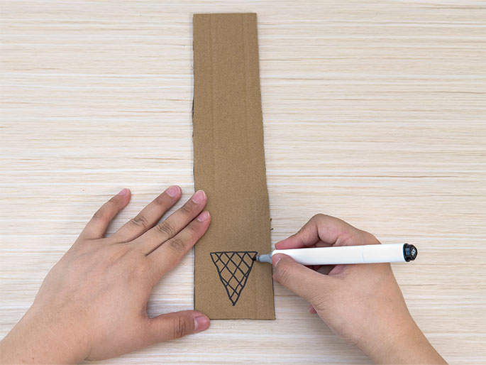 On a light wood-grain surface, a hand uses a white marker to draw an ice cream cone shape onto a vertical strip of brown cardboard. Another hand holds the cardboard steady. This image likely shows a stage in a crafting project where an ice cream cone design is being created on a cardboard base.