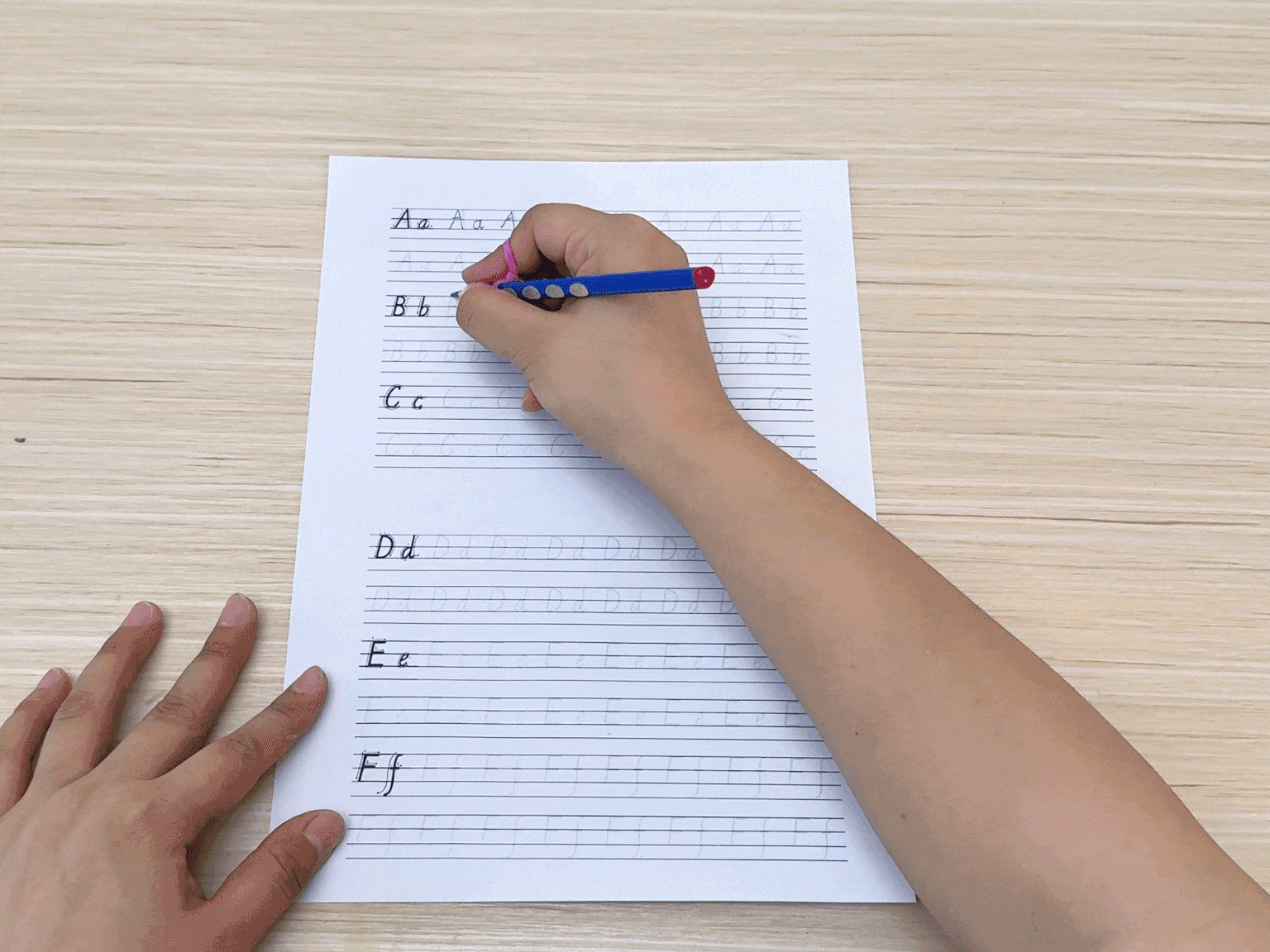 A hand practices cursive writing on lined paper. The letters A through F are shown, with the hand repeatedly writing each letter across the line. This demonstrates a handwriting exercise.