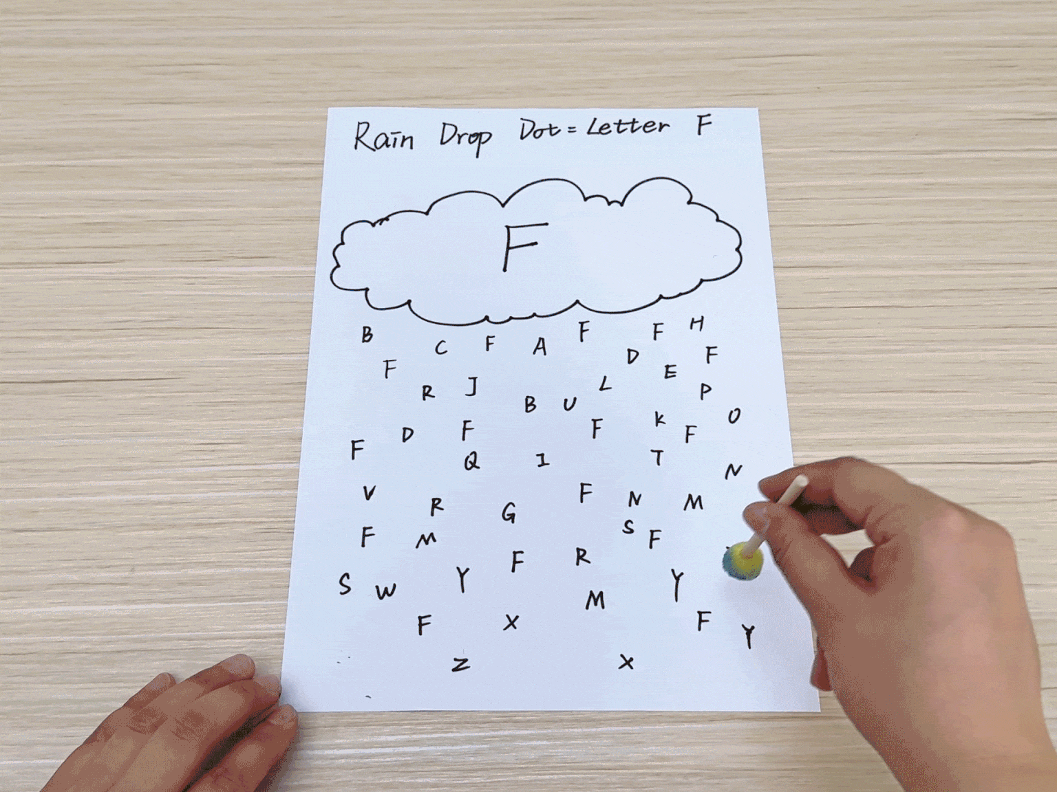 A hand uses a cotton swab to dot paint onto a worksheet titled Rain Drop Dot - Letter F. The worksheet features a cloud with the letter F and a grid of letters below. The hand dots paint on all the F s in the grid, demonstrating a letter recognition activity.