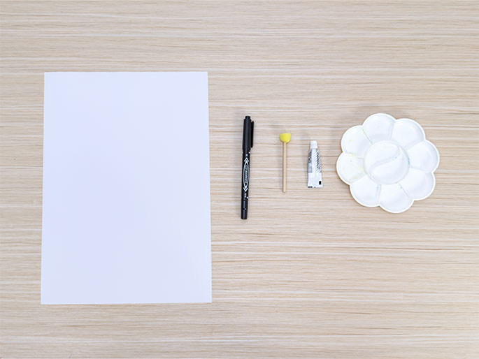 Arranged on a light wood-grain surface are several art supplies: a blank sheet of white paper, a black marker pen, a small wooden stick with a yellow sponge tip, a tube of white glue or paint, and a white flower-shaped paint palette with empty wells. This image depicts a setup for an art or craft project, ready for materials to be added to the palette and used on the paper.