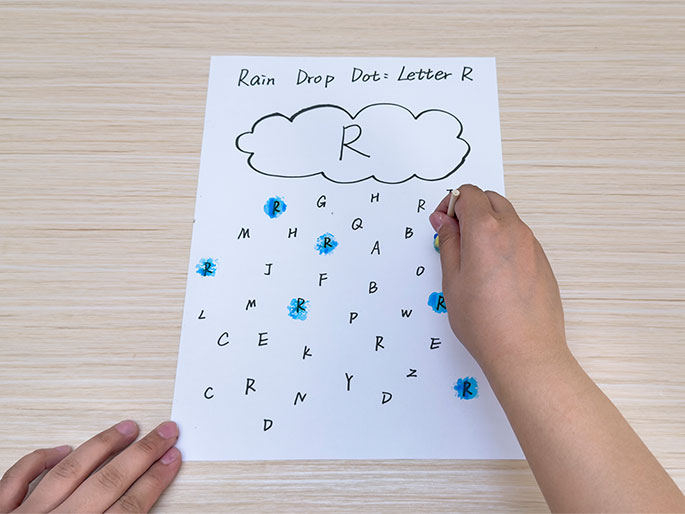 Two hands are interacting with a piece of white paper titled Rain Drop Dot: Letter R on a light wooden table. A hand at the bottom left holds the paper, while a hand on the right uses a small wooden stick with a yellow sponge tip dipped in blue paint to dab the letter R on the paper. A hand-drawn cloud with a large R inside sits at the top of the page. Below the cloud, a grid of letters is scattered, and several  Rs have already been marked with blue dots. This image depicts a child engaged in a letter recognition activity or game.