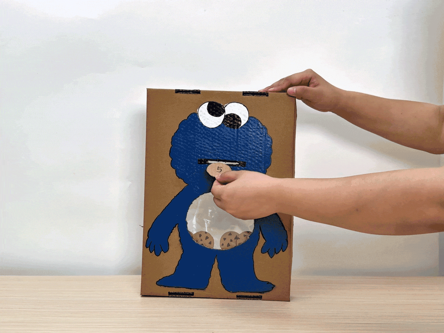 Hands interact with a cardboard cutout of Cookie Monster. The cutout is painted blue with large eyes and a mouth. A clear plastic bag is attached to the belly area, filled with small cookie replicas. A hand pretends to feed Cookie Monster by placing a cookie in its mouth. This playful setup likely serves as a children's activity or game.