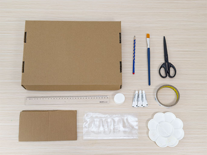 An assortment of craft supplies is laid out on a light wood-grain surface. The items include a flattened cardboard box, a clear plastic ruler, a smaller piece of cardboard, a clear plastic bag, a pencil, a paintbrush, scissors, a roll of clear tape, three small tubes of paint or glue, a small white container with a lid, and a flower-shaped plastic paint palette. The arrangement suggests preparation for a craft or art project.