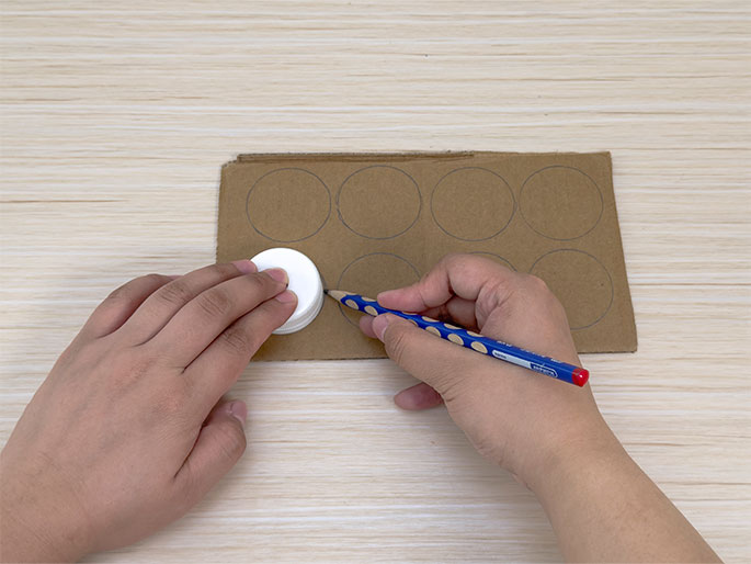 Two hands are shown on a light wood-grain surface, tracing circles onto a piece of brown cardboard. The left hand holds a small, white plastic lid, likely a bottle cap, while the right hand uses a blue and white polka-dotted pencil to trace around the lid. Several, presumably traced in the same manner, are already visible on the cardboard. This image likely circles demonstrates a step in a craft project involving recycled materials.