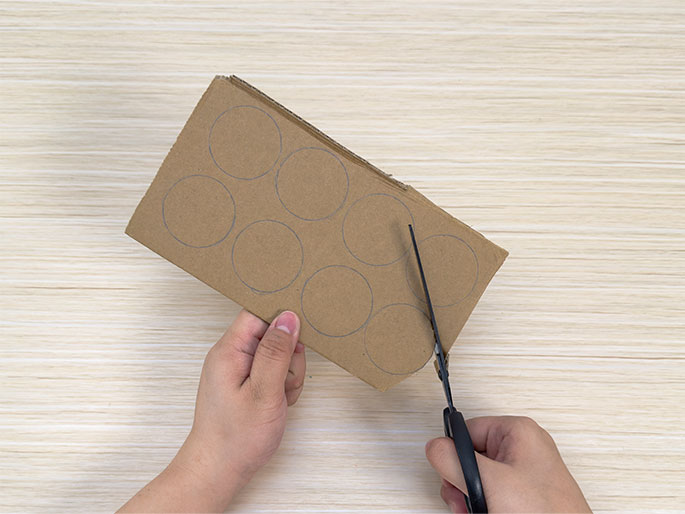 Two hands are shown working with a piece of brown cardboard on a light wood-grained surface. The left hand holds the cardboard, which has several pencil-drawn circles on it. The right hand holds a pair of scissors and is in the process of cutting the cardboard, starting at the edge and moving towards the drawn circles. This image likely depicts a step in a craft project, where circular pieces are being cut out of the cardboard.