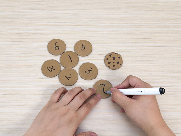 Several small, round cardboard pieces are arranged on a light wood-grain surface. Most of them have numbers written on them, from 1 to 7. One cardboard circle has brown markings drawn on it, resembling a chocolate chip cookie. Two hands are visible ; the left hand steadies a cardboard circle while the right hand uses a white marker to write the number 7 on it. This image seems to depict the creation of a homemade counting or matching game using cardboard cutouts.