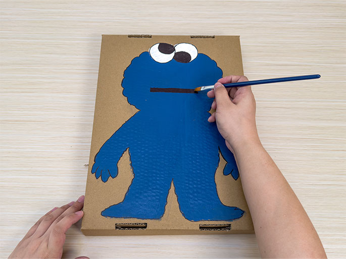 On a light wood-grain surface, a piece of cardboard is being painted. The cardboard has the outline of a figure resembling Cookie Monster from Sesame Street. Two hands are visible; the left hand holds the bottom corner of the cardboard steady, while the right hand uses a paintbrush to apply blue paint within the figure's outline. The figure's eyes are already painted, and a brown stripe, presumably representing Cookie Monster's mouth, has been started. This image captures the process of creating a Cookie Monster painting on cardboard.