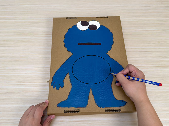 A piece of cardboard, painted blue and shaped like Cookie Monster from Sesame Street, rests on a light wood-grain surface. The Cookie Monster figure has white and dark brown eyes and a dark brown mouth. Two hands are interacting with the cardboard; the The left hand holds the bottom left corner, while the right hand uses a blue and white polka-dotted pencil to draw a circle on the figure's belly. This image likely depicts a stage in a craft project involving creating a Cookie Monster figure.