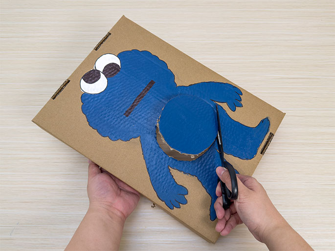 Two hands are working with a piece of cardboard on a light wood-grained surface. The cardboard has a painted blue Cookie Monster figure on it. A circular section of the Cookie Monster's belly has been cut out partially. The left hand holds the cardboard, while the right hand uses scissors to continue cutting out the circular portion. This image likely shows a step in creating a Cookie Monster craft, possibly a feeding game or a puppet.