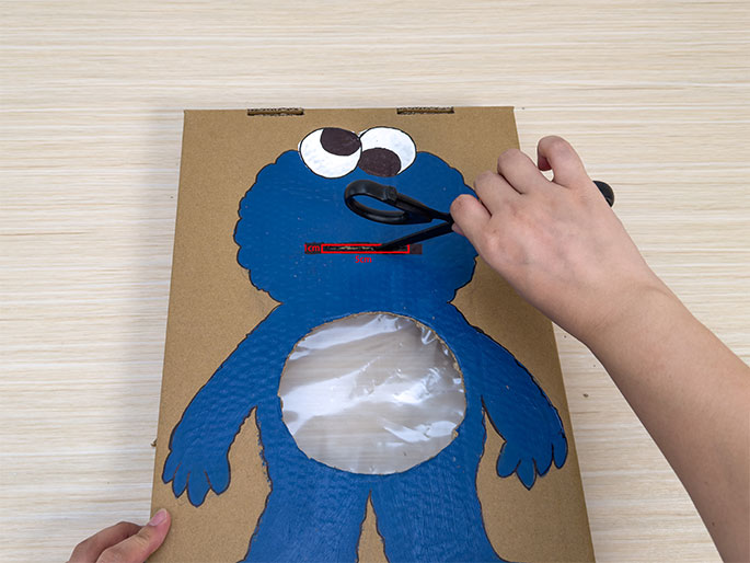 On a light wood-grain surface, a cardboard cutout of Cookie Monster is being modified. The Cookie Monster is painted blue, with a circular opening cut out of its belly, now covered with a clear plastic sheet. Two hands are visible; the left hand holds the cardboard while the right hand uses scissors to cut a slit in the Cookie Monster's mouth area. This image seems to depict the creation of a Cookie Monster feeding game or toy from cardboard.