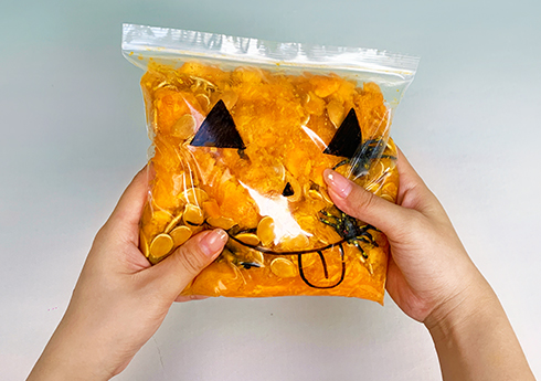 Two hands gently hold a clear plastic bag filled with pumpkin-coloured material, possibly pumpkin puree or a similar substance. Black marker pen details create a playful jack-o'-lantern face on the bag, and small black objects, possibly plastic spiders, are visible within. The overall effect is a fun, slightly spooky, and visually appealing Halloween-themed sensory item. The image is bright and well-lit, highlighting the playful design.