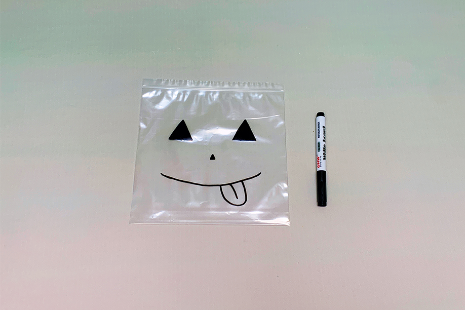 A clear plastic bag, decorated with a simple black marker pen drawing of a playful jack-o'-lantern face, lies on a light beige surface. The face features triangular eyes, a small nose, a curved mouth, and a protruding tongue. Beside the bag sits a black permanent marker pen, suggesting the tool used to create the design. The overall impression is of a simple, fun, and readily achievable Halloween craft project. The background is a softly lit, even tone.