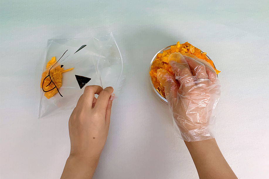 Two hands are shown preparing a Halloween craft project. One hand holds a clear plastic bag, already partially decorated with a black marker pen to resemble a jack-o'-lantern face, whilst the other hand, wearing a clear plastic glove, scoops bright orange, shredded material (likely pumpkin) from a small bowl. The scene is set against a light, off-white background, with the focus on the careful transfer of the orange material into the bag. The image depicts a step in creating a sensory Halloween activity.