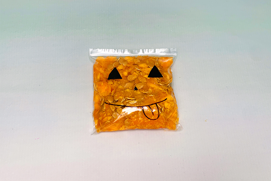 A sealed, clear plastic bag lies on a light grey surface. Inside, a mass of orange material, possibly pumpkin puree or a similar substance, is visible. Black marker pen markings on the bag form a simple, playful jack-o'-lantern face, complete with triangular eyes, a small nose, and a long, protruding tongue. The bag's contents appear slightly translucent, and some small, light-coloured objects are interspersed within the orange material. The overall impression is of a completed Halloween-themed sensory item, ready for use.