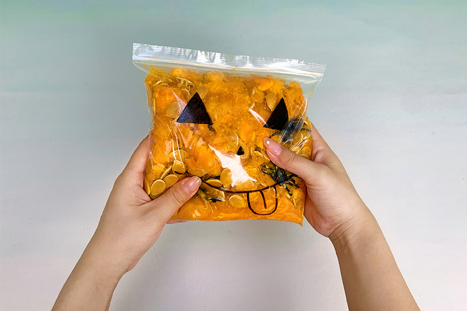 Two hands gently cradle a clear plastic bag filled with a vibrant orange substance, possibly pumpkin purée or a similar material. Black marker pen details create a whimsical jack-o'-lantern face on the bag, with triangular eyes, a simple nose, and a playful, protruding tongue. Small, dark objects, possibly plastic spiders, are visible within the orange mixture. The overall effect is a fun, slightly spooky, and visually appealing Halloween-themed sensory item. The image is brightly lit, focusing on the playful design and the hands holding the bag.