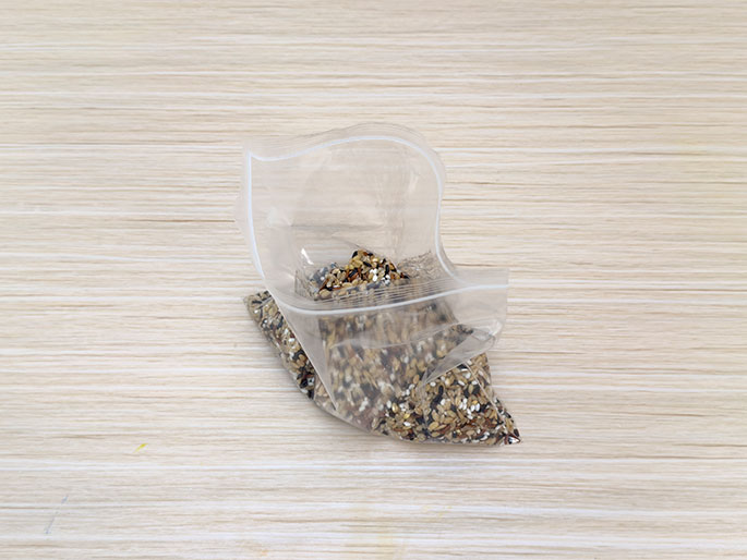 A clear plastic zip-top bag partially filled with mixed rice grains rests on a light wood-grain surface. The bag is sealed and lies at an angle, with the top portion folded over. The mixed rice inside is visible through the plastic. This image likely depicts a step in a DIY or sensory activity using rice.
