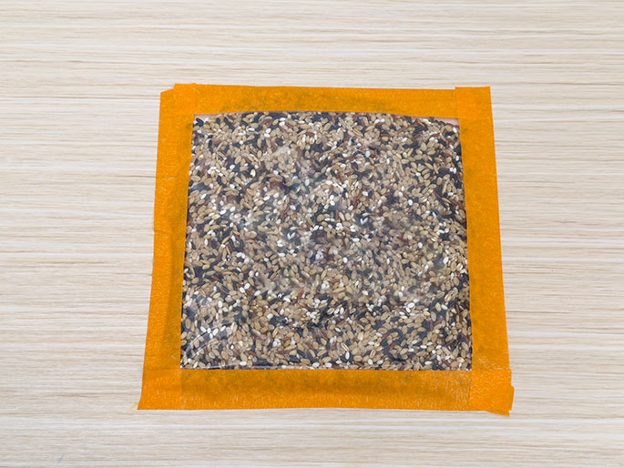 On a light wood-grain surface, a clear plastic bag filled with mixed rice grains is shown. The bag is sealed and reinforced with orange tape around all four edges. This image likely illustrates a step in creating a sensory toy or activity, where the tape adds durability and a visual border.