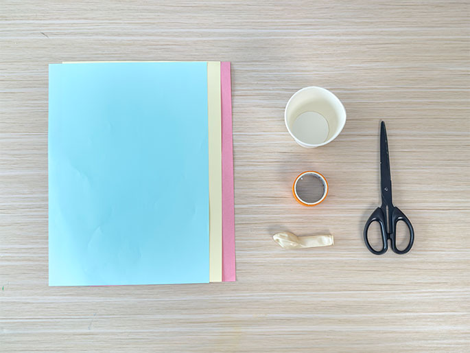 Arranged on a light wood-grain surface are various craft supplies. On the left, a stack of pastel-colored paper sheets in light blue, yellow, and pink is visible. To the right, a small white paper cup sits next to a roll of orange tape. A deflated, pale yellow balloon and a pair of black scissors complete the arrangement. The items suggest preparation for a children's craft or science activity.
