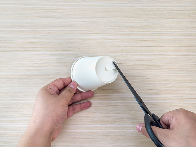 Two hands are shown manipulating a small white paper cup and a pair of black scissors on a light wood-grain table. One hand holds the cup on its side while the other hand uses the scissors to make a small cut in the rim of the cup. This image likely depicts a step in a DIY or craft project involving the paper cup.