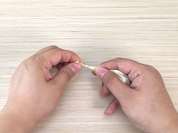 Two hands are shown tying a knot in the neck of a deflated, pale yellow balloon. The hands are positioned against a light wood-grain background. This image illustrates the process of preparing a balloon for inflation, likely for a craft or science activity.
