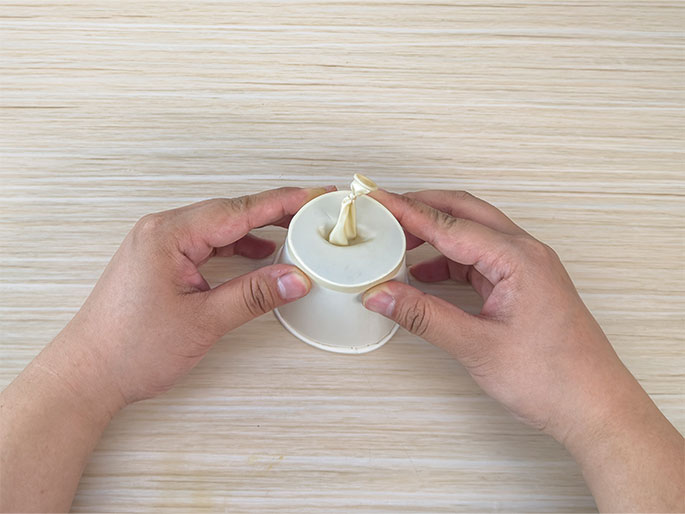 Two hands hold a small white paper cup on a light wood-grain surface. The knotted neck of a pale yellow balloon is stretched over the opening of the cup, with the deflated balloon body draped inside. This image appears to demonstrate a stage in a science experiment or craft activity, possibly involving sound or air pressure.