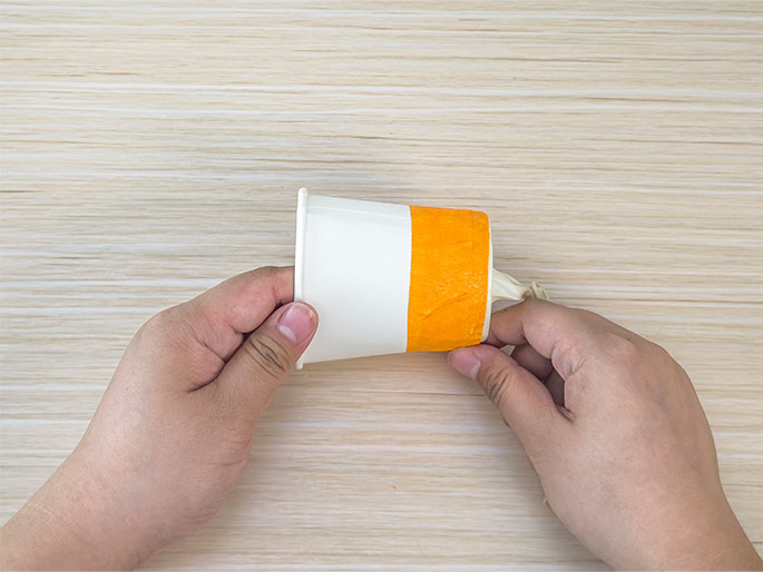 Two hands hold a small white paper cup on its side against a light wood-grain background. A band of orange tape secures the knotted end of a pale yellow balloon to the cup. The balloon itself is deflated and tucked inside the cup. This image likely depicts a completed step in a DIY project or science experiment.