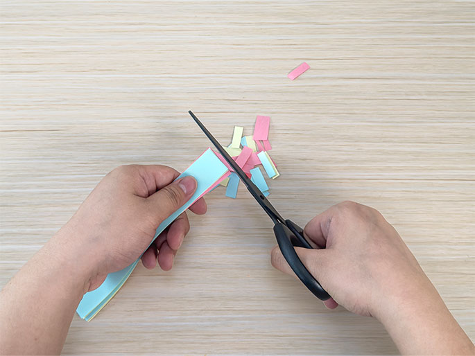Two hands are engaged in a paper-cutting activity on a light wood-grain table. One hand holds a stack of thin strips of pastel-colored paper – light blue, pink, and yellow – while the other hand uses black scissors to cut the strips. Several small pieces of cut paper lie scattered near the scissors, indicating ongoing progress in the activity. This image likely depicts a step in a craft project.