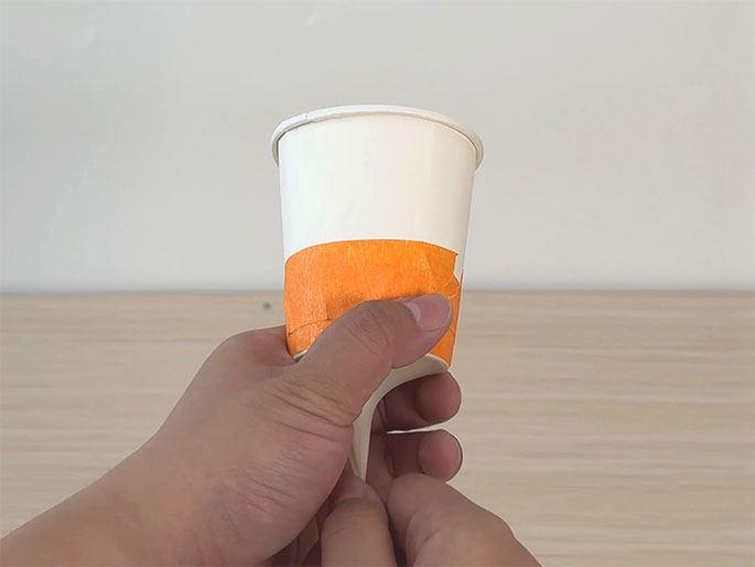 A hand holds a small white paper cup against a plain off-white background. A strip of bright orange tape encircles the cup. A light wood-grain surface is visible in the lower portion of the image. The focus is on the cup, suggesting it's being prepared for a craft or experiment.