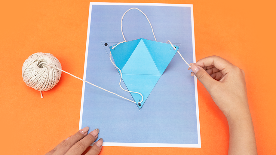 Two hands thread string through the holes of a light blue paper tetrahedron resting on a lavender piece of paper, which sits on an orange background; a ball of string is visible to the left.