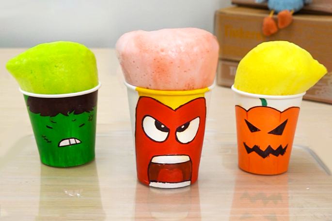 Three paper cups overflow with colorful foam. From left to right: a green cup with a Hulk face and bright green foam, a red cup with an angry face and pink foam, and an orange cup with a jack-o'-lantern face and yellow foam. They sit on a light-colored surface. This image likely depicts the results of a science experiment or a fun children's activity.