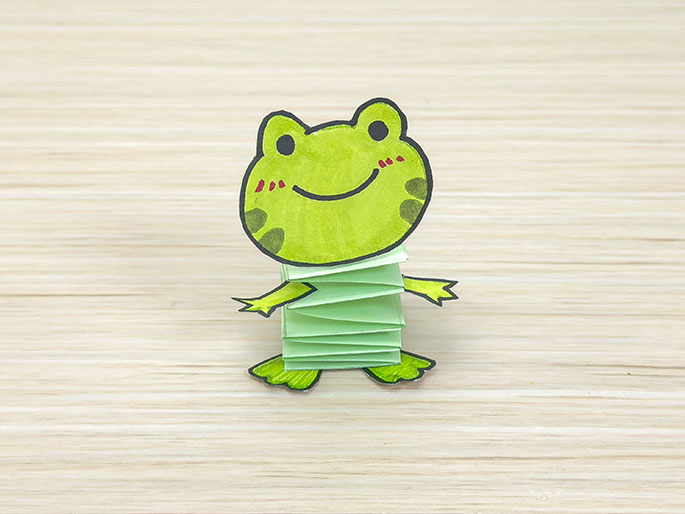 A completed paper craft frog sits on a light wood surface. The frog's head is a drawing on light green paper, while its body is formed from a folded, accordion-style stack of pale green paper. Small, drawn arms and feet complete the figure.