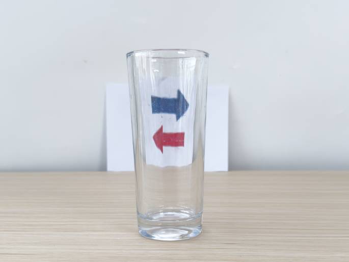 A clear glass sits on a light wooden table with a piece of white paper positioned behind it. The paper features two arrows: a blue arrow pointing to the right and a red arrow pointing to the left. A hand pours water from a clear glass pitcher into the empty glass. As the glass fills, the arrows appear to change direction due to the refraction of light through the water. This video demonstrates a simple science experiment illustrating the principles of light refraction.