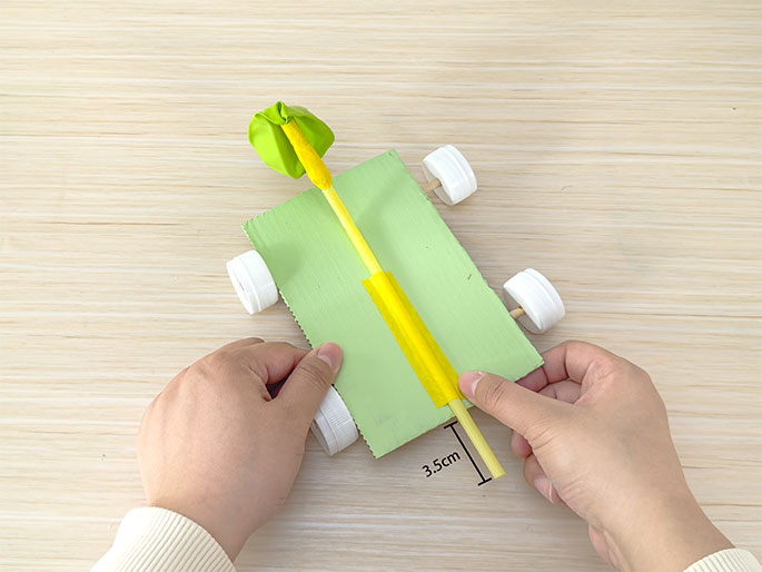 Two hands hold a small, light green cardboard car on a light woodgrain surface. The car has four white bottle cap wheels attached with wooden dowels as axles. A yellow straw is taped to the top of the car, and a deflated lime green balloon is attached to the other end of the straw. The straw extends off the back of the car. A measurement line indicates the straw extends 3.5cm beyond the back edge of the cardboard.