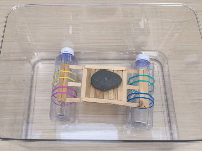 Hands hold a simple raft or boat constructed from recycled materials against a light wood background. The boat's base is a platform made of craft sticks glued together. Two clear plastic bottles are attached to the underside of the platform, one on each end. Craft sticks are fastened to the bottles with colorful rubber bands—pink and yellow on one bottle, and teal and blue on the other. One hand holds the boat while the other adjusts a rubber band. This image likely depicts a children's craft project or a simple science experiment demonstrating buoyancy.