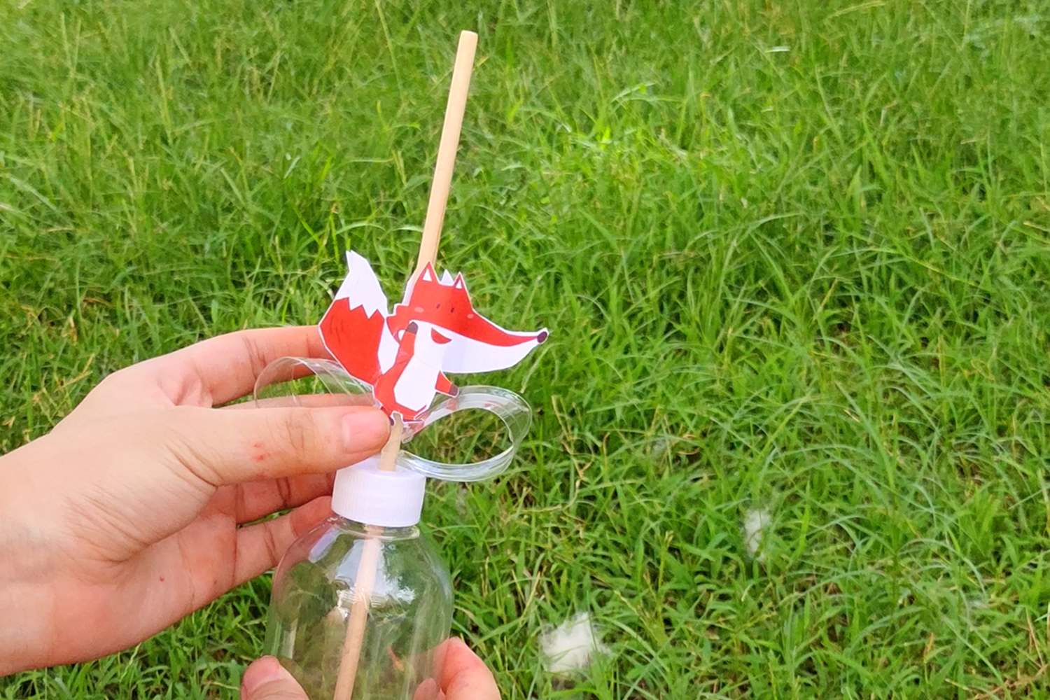 Two hands hold a clear plastic bottle with a wooden stick pushed through the cap and a paper fox attached to the stick; the bottle is outdoors on a patch of grass.