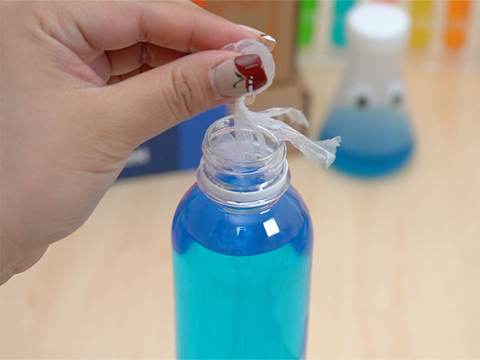 A hand places a crumpled piece of clear plastic sheeting into the opening of a clear plastic bottle containing blue liquid. The bottle cap is held between the fingers of the hand inserting the plastic. In the background, a blurred, decorated beaker with googly eyes and blue liquid is visible. The items are on a light wood-grain surface.
