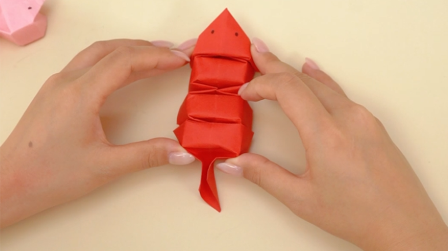 Two hands hold a completed red origami mouse, its body made of folded segments and a small, pointed tail. A small, pink origami object is visible in the top left corner.