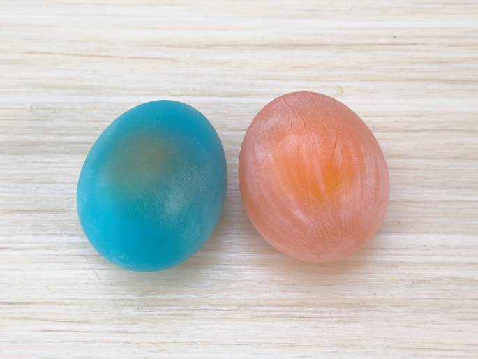Two dyed eggs rest on a light woodgrain surface. The egg on the left is a vibrant teal or turquoise, while the egg on the right is a light peach or coral color. Both eggs appear slightly translucent, revealing a hint of the yolk inside. The image showcases the results of an egg dyeing process.