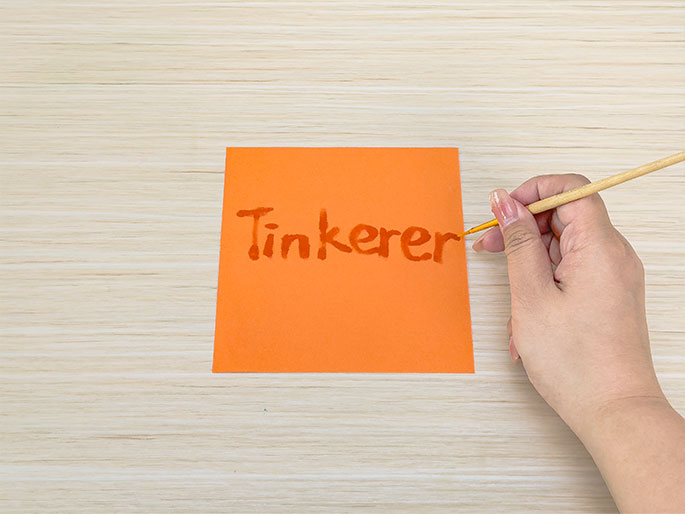 On a light-colored woodgrain surface, a hand holds a small paintbrush and writes the word Tinkerer in orange paint on a bright orange square of paper. The hand is in the process of completing the last letter. This image likely represents a craft activity or DIY project.