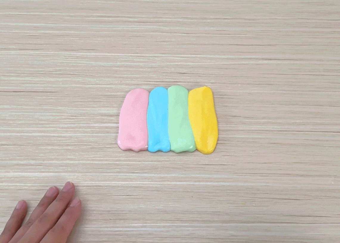 Two hands press down on four parallel lines of pastel-colored slime or playdough on a light wooden surface. The colors are pink, light purple, light green, and yellow. The hands then gather the slime/playdough together and stretch it, revealing the swirling pastel colors inside. The video demonstrates the playful manipulation of a colorful, stretchy substance, likely slime or a similar material.