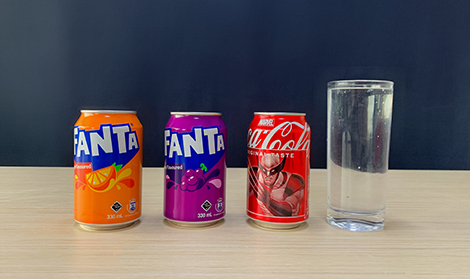Three cans of fizzy drinks – Fanta Orange, Fanta Grape, and Coca-Cola – stand on a table next to a tall glass of water, all against a dark blue background.