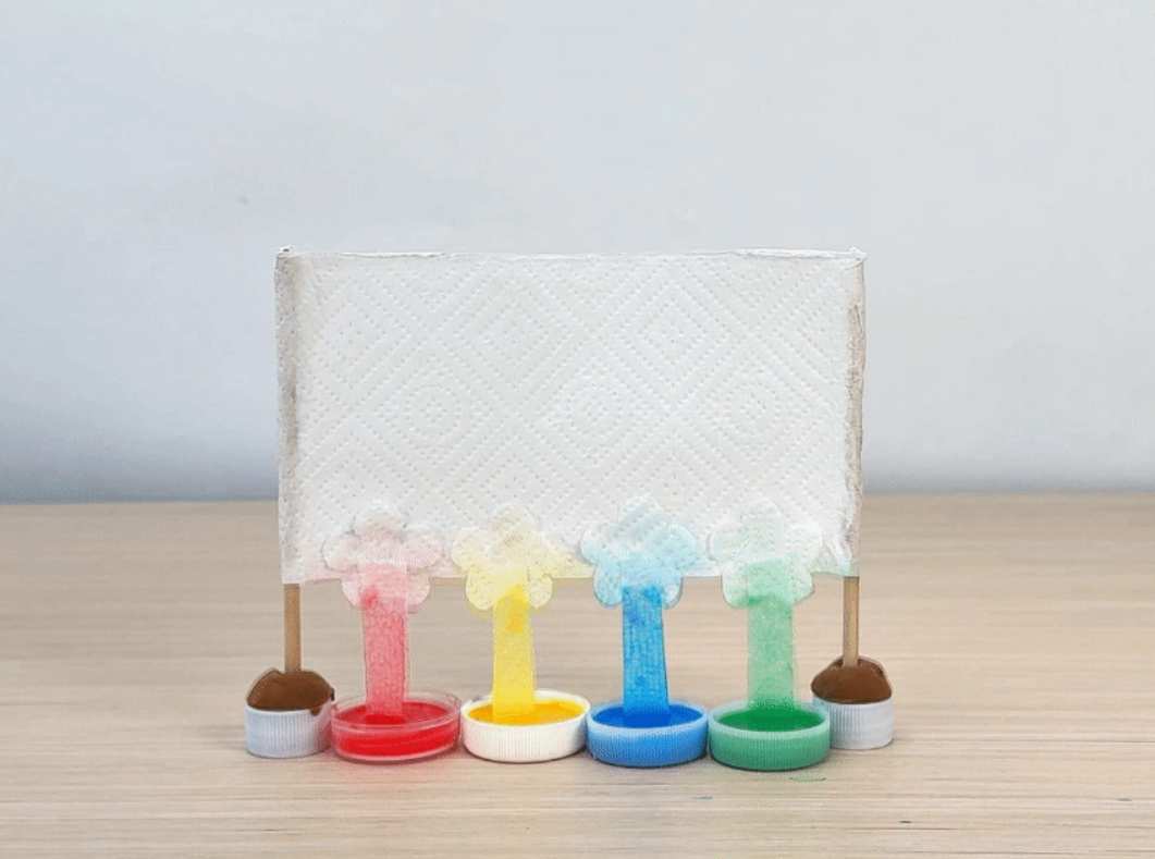 A time-lapse video demonstrates a color mixing experiment using paper towels and colored water. Four small, clear plastic bottle caps containing red, yellow, orange, and green water are arranged in a row on a light wooden surface. Small wooden sticks are inserted into modeling clay bases and placed behind each cap. A white paper towel is draped over the sticks, with the bottom edge submerged in the colored water. As time passes, the colored water wicks up the paper towel via capillary action, creating vertical streaks of color that gradually blend together where they meet. The video focuses on the process of the colors traveling up the paper towel and mixing.